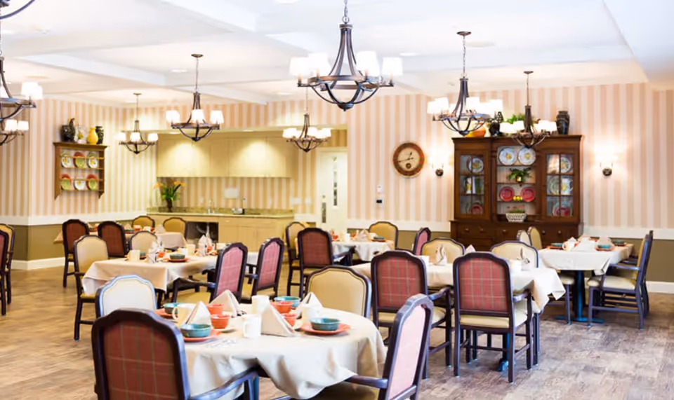 A bright dining room with multiple tables set with dishes, upholstered chairs, chandeliers, and a china cabinet against a striped wall.