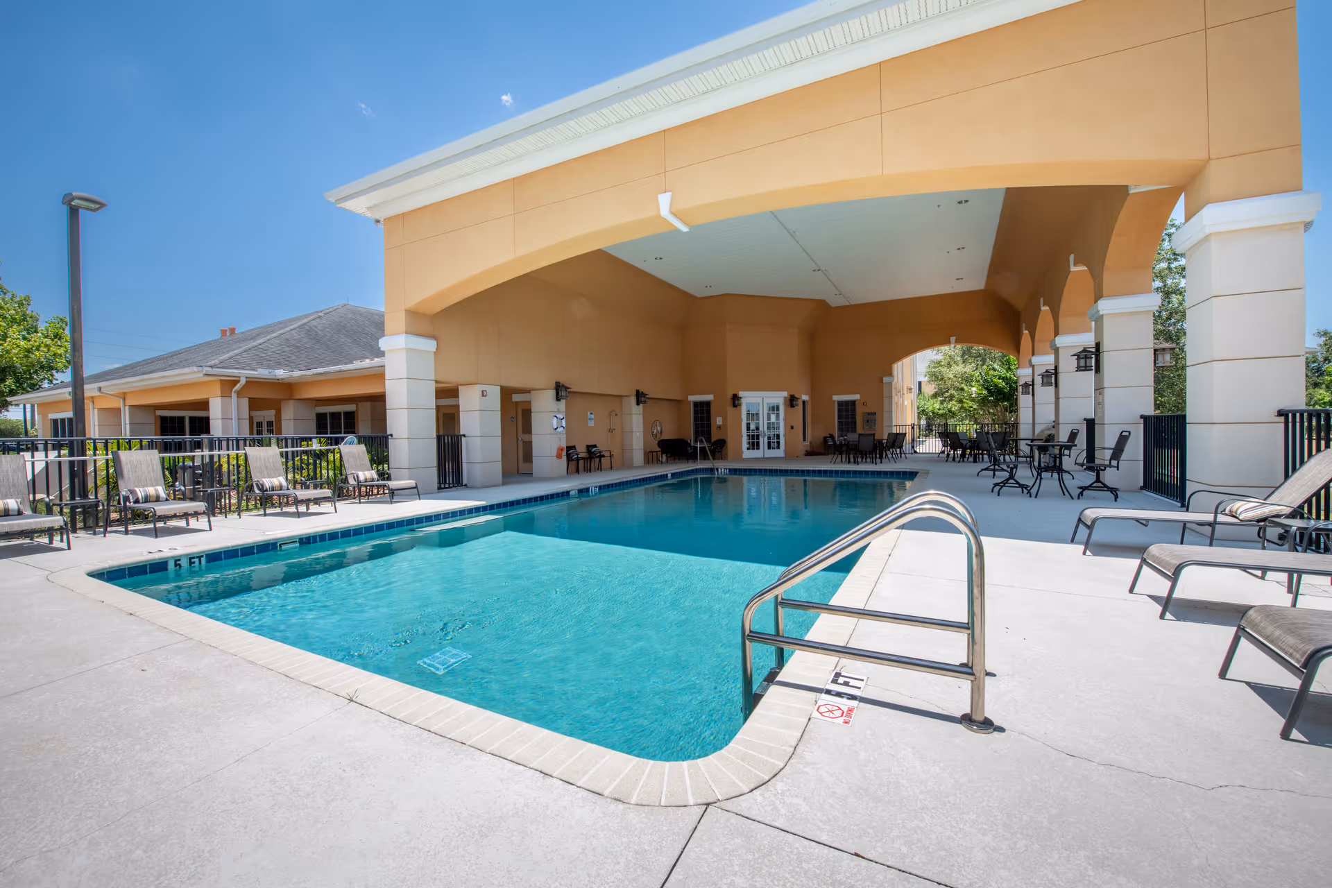 Outdoor swimming pool area at Sweet Water Grand The Bridges Assisted Living with lounge chairs and tables under a covered patio and clear blue sky.