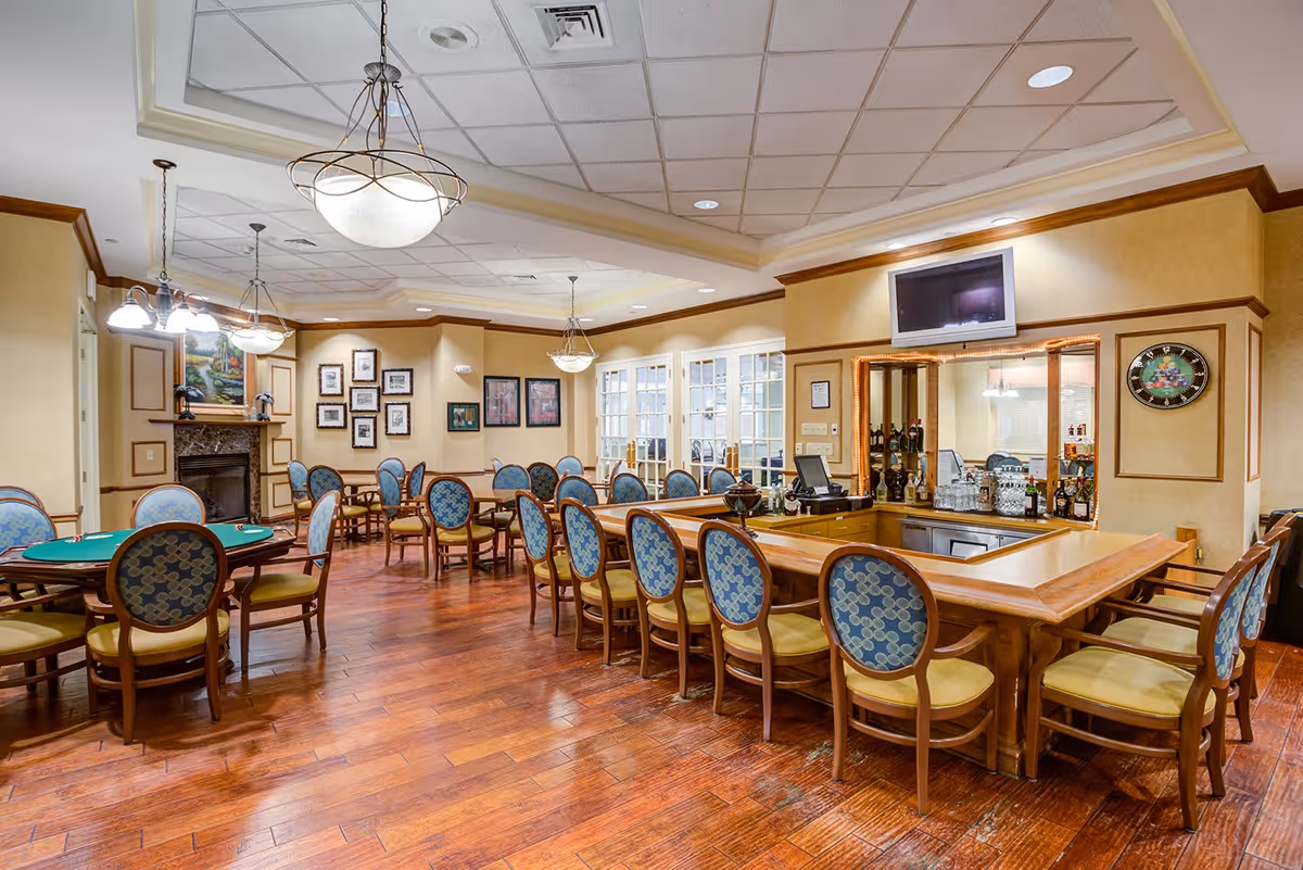 A spacious and well-lit dining and lounge area with wooden floors, multiple round tables surrounded by blue and yellow cushioned chairs, a wooden bar with high chairs, a wall-mounted TV, and framed artwork on the walls. The ceiling has recessed lighting and hanging light fixtures.