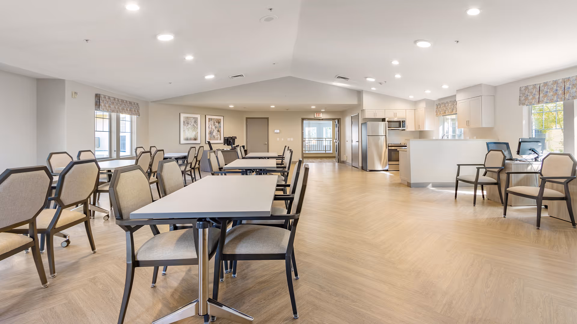 Bright communal dining and activity room with multiple tables and chairs and a kitchenette in the background.