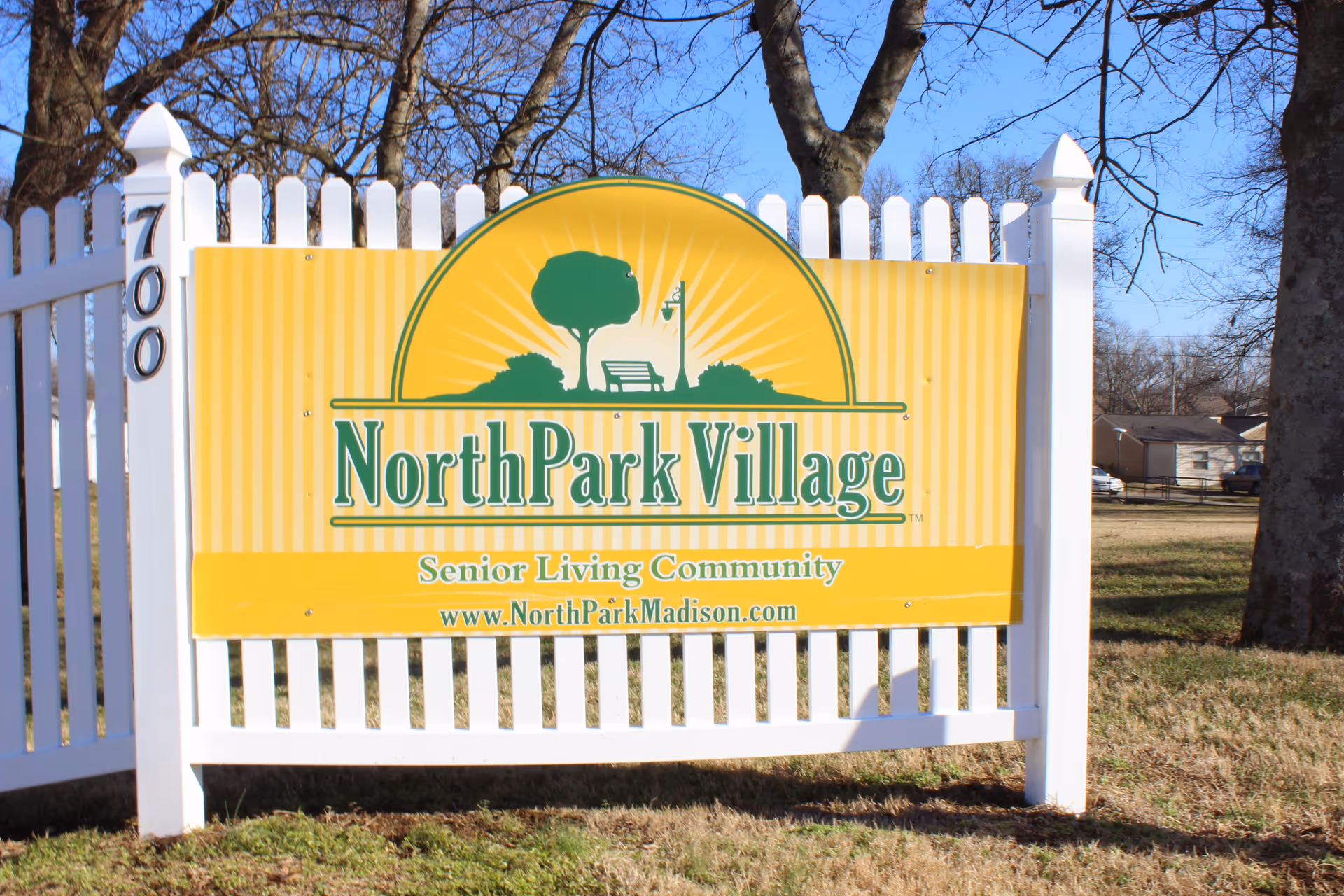 A yellow sign with green text that reads 'North Park Village Senior Living Community www.NorthParkMadison.com' mounted on a white picket fence with the number 700 on the left post, set outdoors with trees and grass visible in the background.