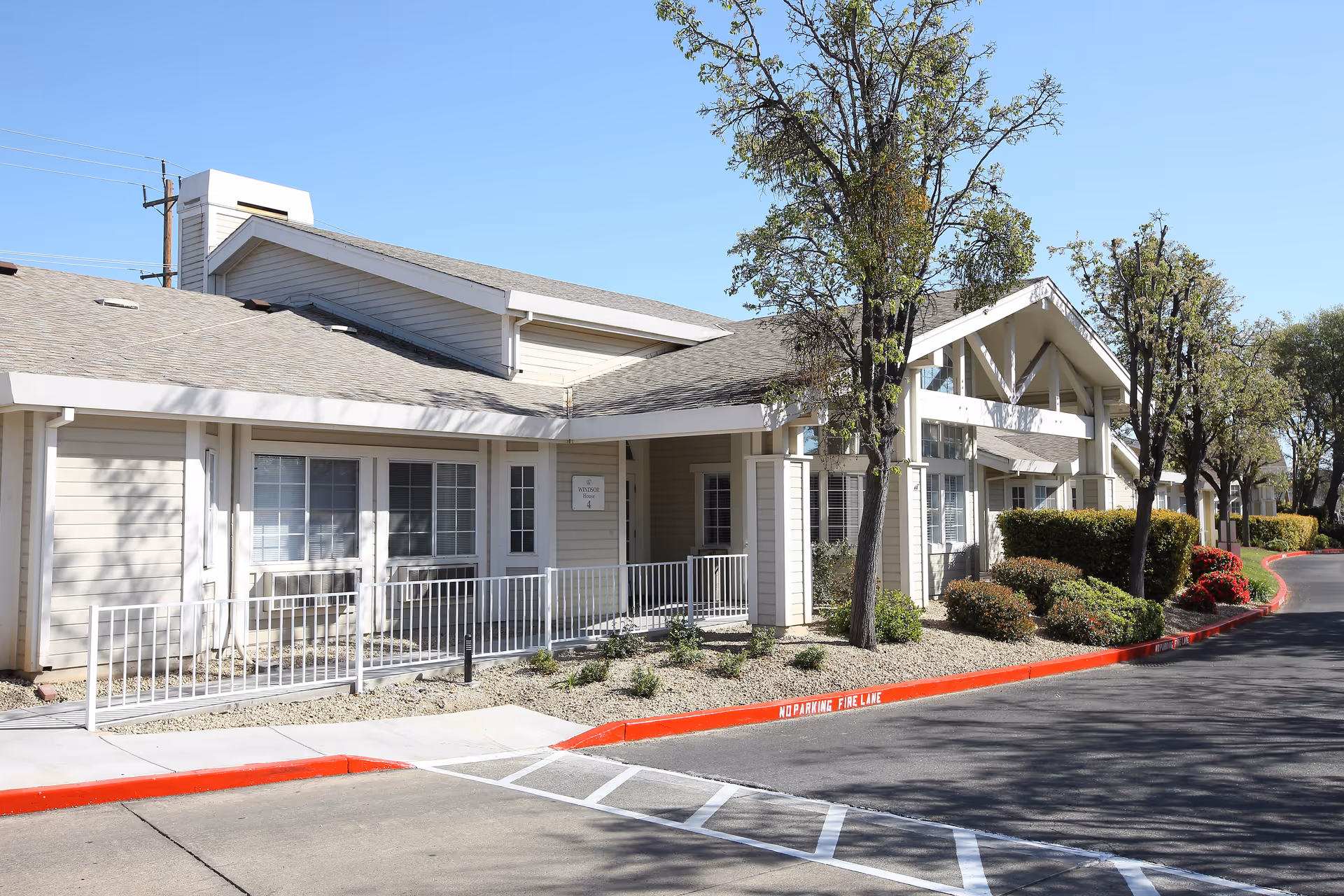 Exterior view of a single-story building with beige siding and a gray shingled roof, surrounded by small trees and shrubs. There is a sidewalk with a white railing and a red curb marked 'NO PARKING FIRE LANE' in front of the building under a clear blue sky.