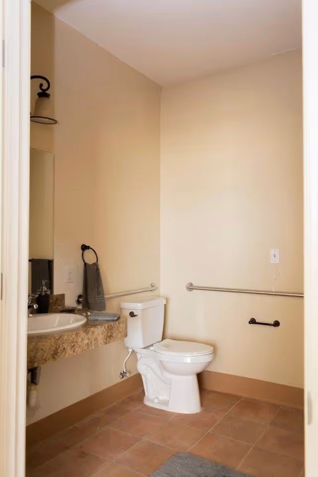 Accessible bathroom with a toilet, sink set in a stone countertop, grab bars on the walls, and beige tiled floor and walls.
