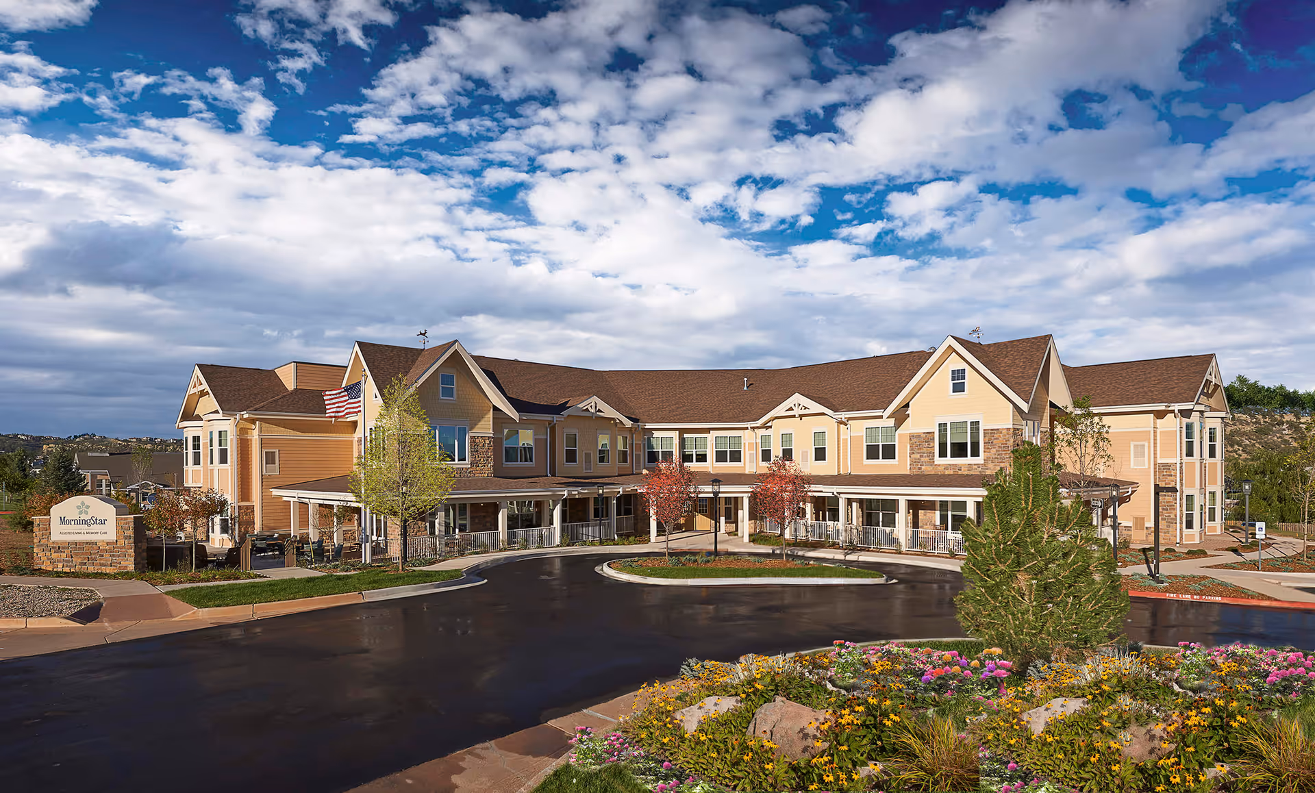 Exterior view of MorningStar Assisted Living & Memory Care at Mountain Shadows, a two-story building with beige and brown siding, multiple windows, and a covered entrance. The building is surrounded by a circular driveway, landscaped greenery, colorful flowers, and trees under a partly cloudy blue sky.