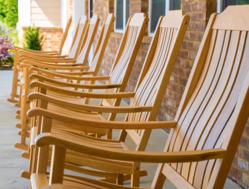 A row of wooden rocking chairs lined up on a covered porch with a brick wall and windows in the background.