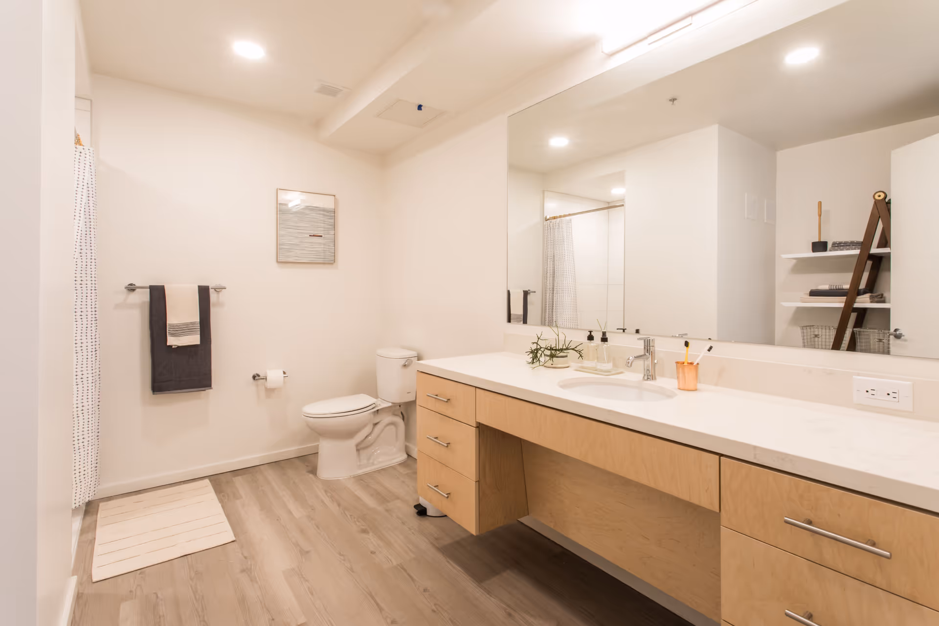 A modern bathroom with light wood flooring and white walls. The room features a white toilet, a large vanity with a white countertop and built-in sink, and a large mirror above the vanity. On the countertop, there are soap dispensers, a small plant, and a toothbrush holder. A towel rack with two towels is mounted on the wall next to the toilet. A shower with a white curtain is partially visible on the left side. There are shelves with towels and baskets on the right side of the image.