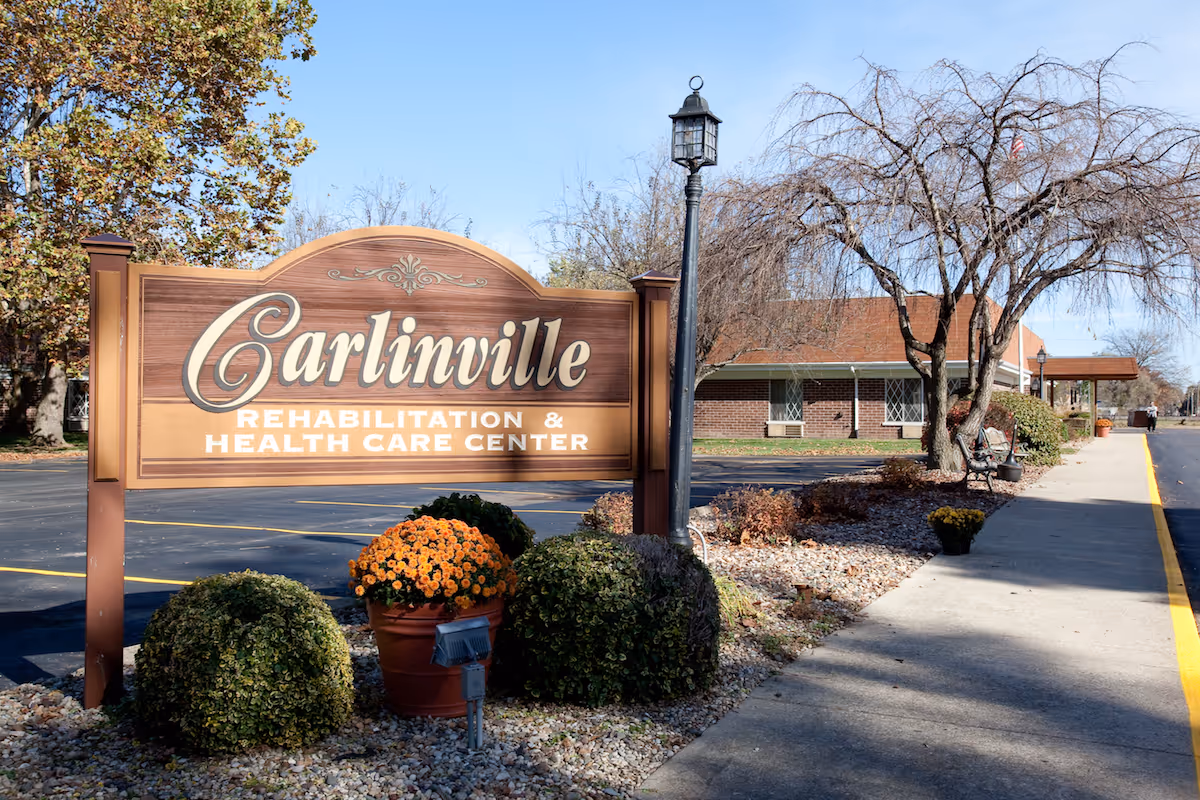 Outdoor view of Carlinville Rehabilitation & Health Care Center sign with bushes and flowers in front, a sidewalk, a lamp post, and part of the building with trees in the background under a clear blue sky.