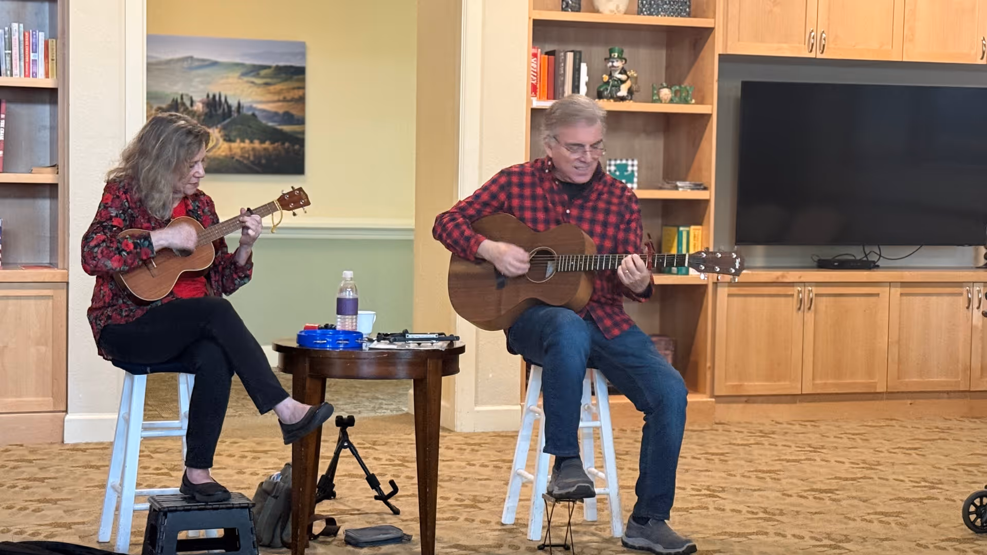 A man and a woman sitting on stools in a living room area playing string instruments, the woman playing a ukulele and the man playing an acoustic guitar. They are positioned near a small round table with a water bottle and some items on it. Behind them are wooden cabinets, shelves with books and decorations, and a large flat-screen TV.