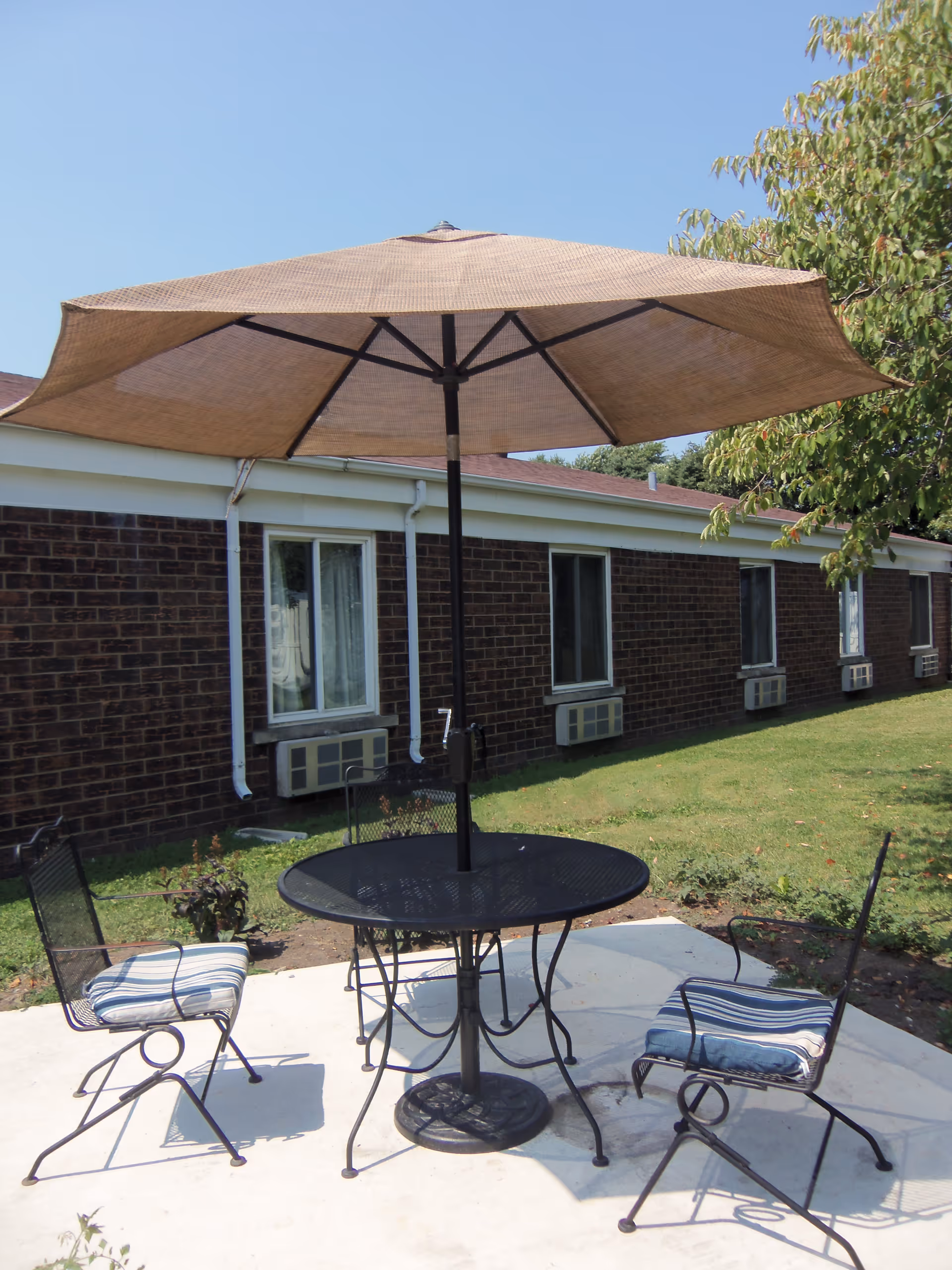 Outdoor patio area with a round black metal table and three matching chairs with striped cushions. A large beige umbrella is open above the table providing shade. The patio is adjacent to a brick building with several windows and air conditioning units. There is green grass and a tree nearby under a clear blue sky.