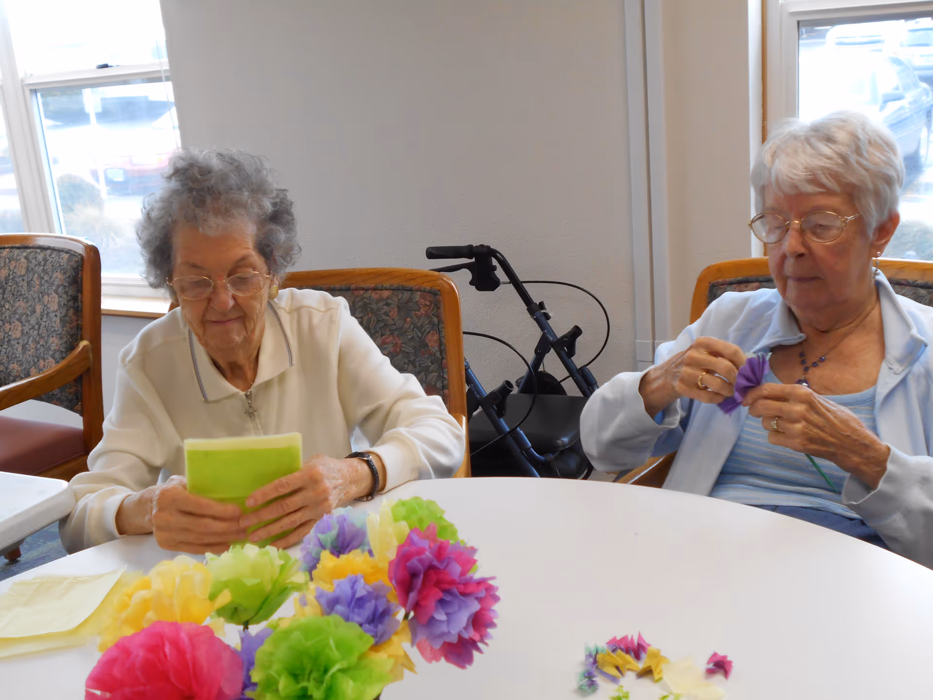 Two elderly women sitting at a round table indoors, making colorful paper flowers. One woman is holding green paper, while the other is shaping a purple flower. A walker is visible behind them near the window.