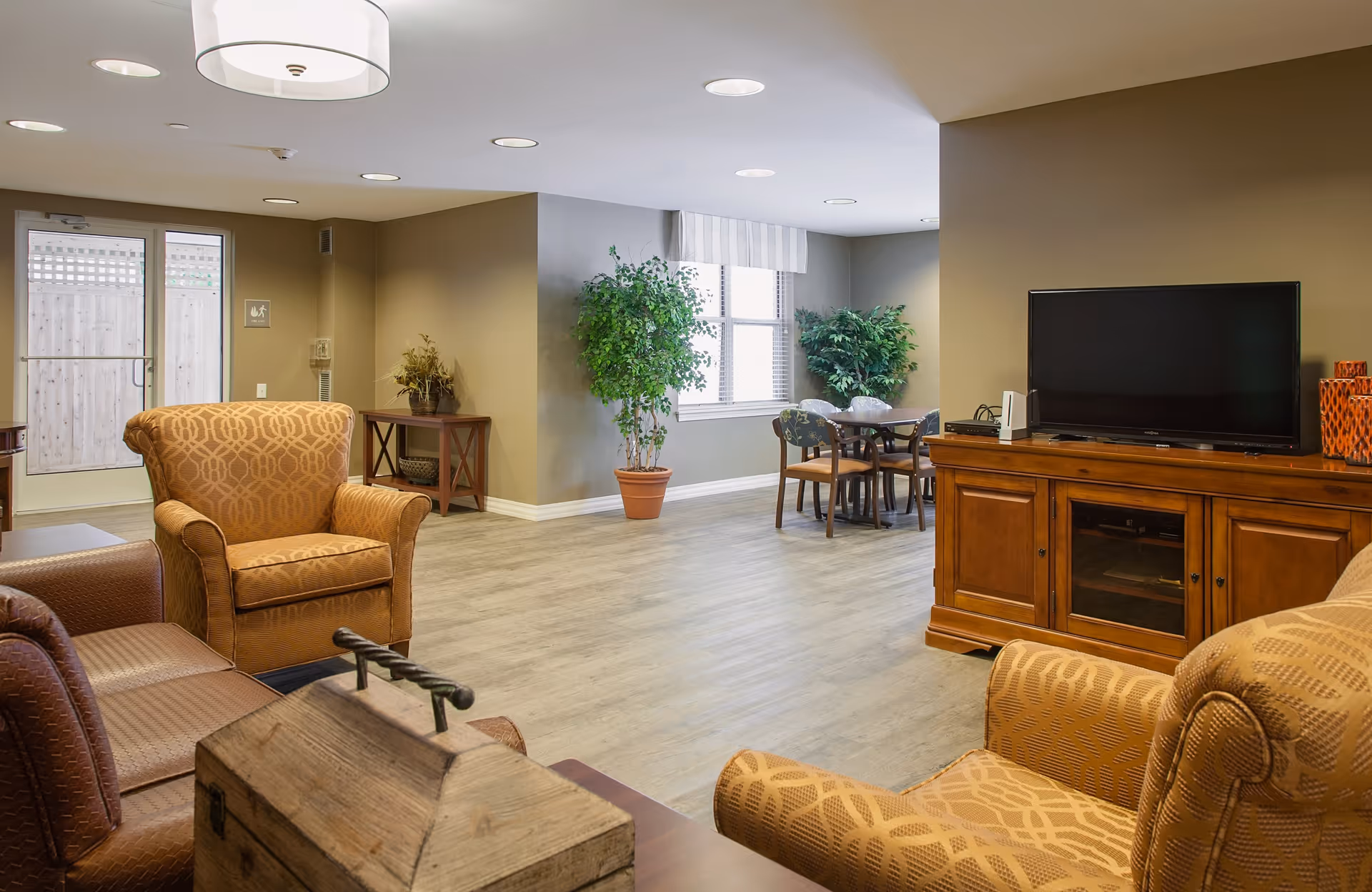 A cozy living room area with patterned armchairs and a brown leather sofa. There is a wooden TV stand with a flat-screen television on top. In the background, there is a small dining table with four chairs near a window with white blinds. The room has light wood flooring, beige walls, and potted plants adding greenery. A glass door leads outside.