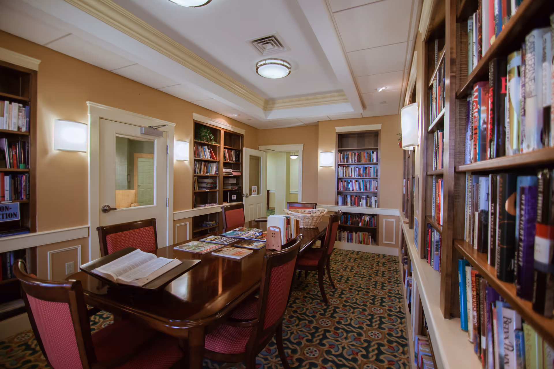 Interior view of a library or reading room with wooden bookshelves filled with books on both sides. A long wooden table with red cushioned chairs is in the center, holding open books and magazines. The room has beige walls, decorative molding, and patterned carpet flooring. Ceiling lights and wall sconces provide illumination.