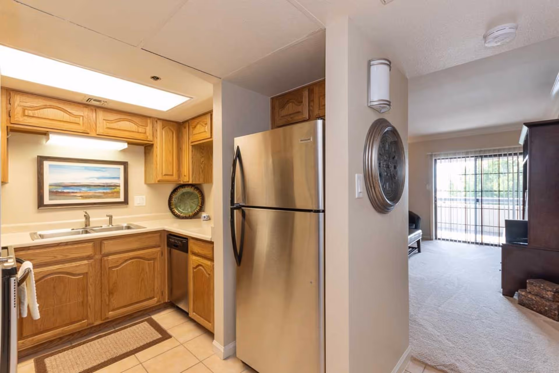 Interior view of a kitchen with wooden cabinets, a stainless steel refrigerator, a double sink, and a dishwasher. The kitchen opens into a living room area with carpeted flooring and a sliding glass door leading to an outdoor balcony.