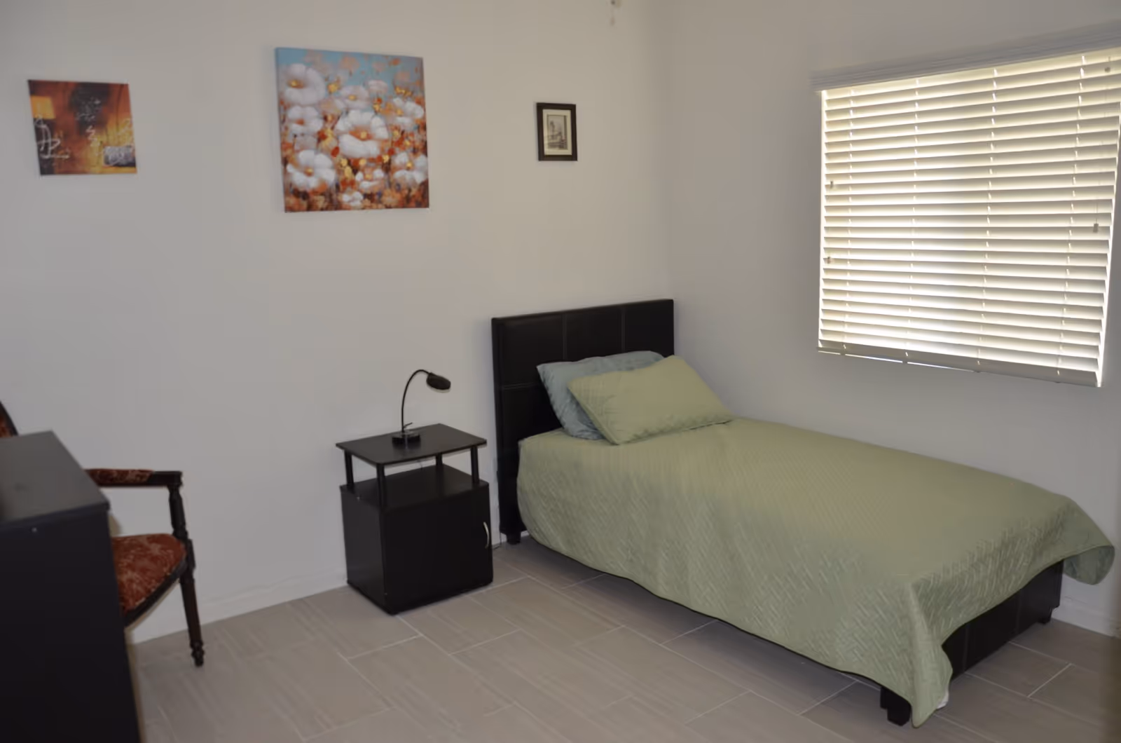 A simple bedroom with a single bed covered with a green quilt and two pillows. Next to the bed is a small black nightstand with a small lamp on it. There is a wooden chair with a red cushion on the left side. The walls are white and decorated with three framed pictures. A window with white blinds is on the right wall.