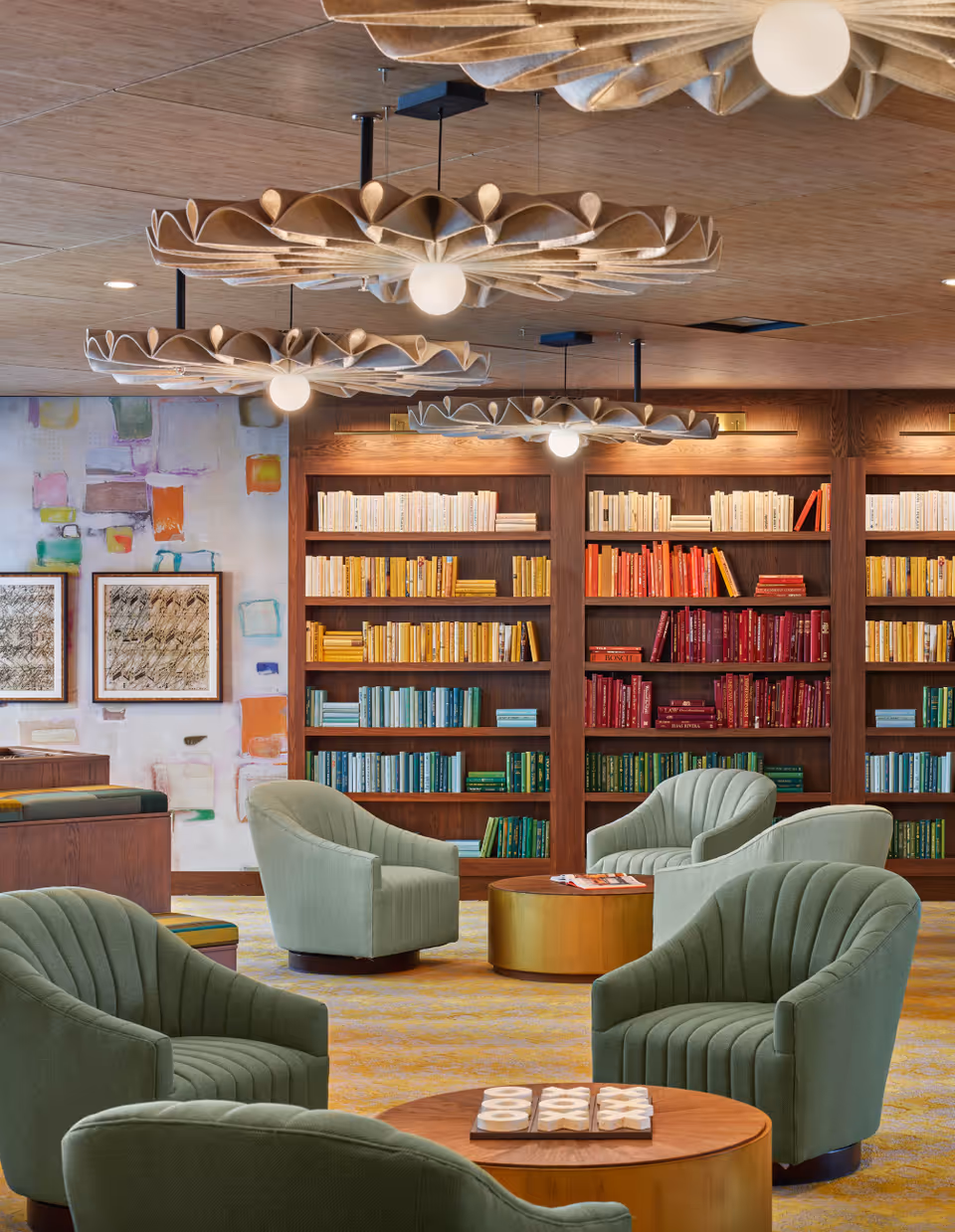 A cozy lounge with green upholstered armchairs, round wooden tables, decorative pendant lights, and bookshelves filled with color-organized books.