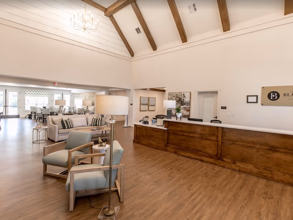 Spacious senior living facility lobby with high vaulted ceiling featuring wooden beams, a wooden reception desk, two light blue armchairs with wooden frames, a beige sofa with striped pillows, floor lamps, and a dining area with tables and chairs in the background.