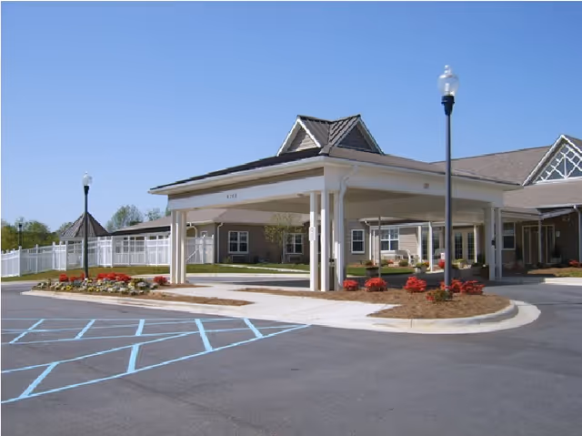 Exterior view of Davidson Health and Rehab Center showing a covered entrance with white pillars, a driveway with marked parking spaces including handicapped spots, landscaped flower beds with red flowers, and a clear blue sky.