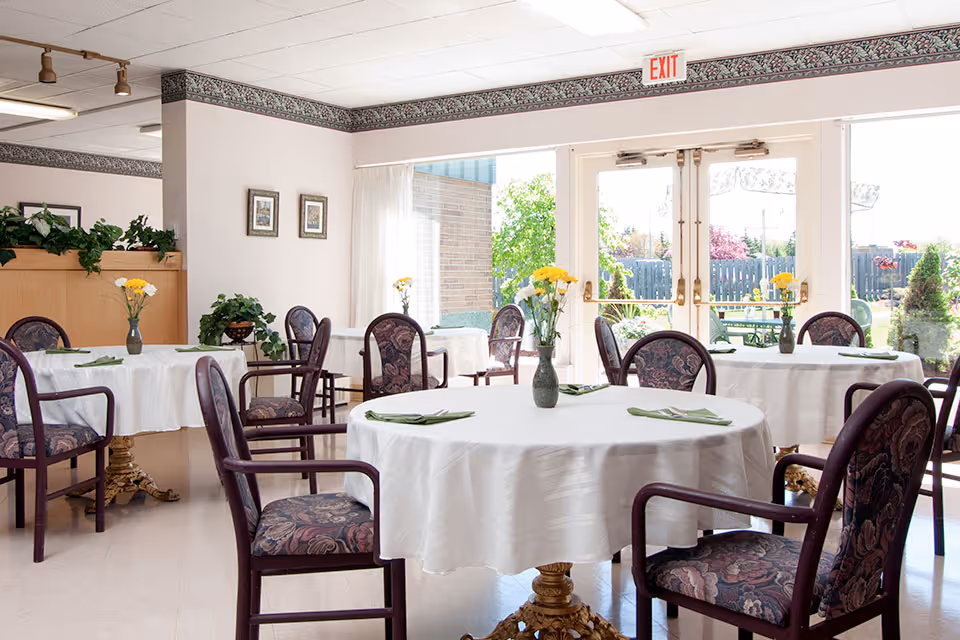Bright dining room with round tables covered in white tablecloths, each set with green napkins and a vase of yellow flowers. Upholstered chairs with floral patterns surround the tables. Large windows and glass doors let in natural light and offer a view of a garden outside.