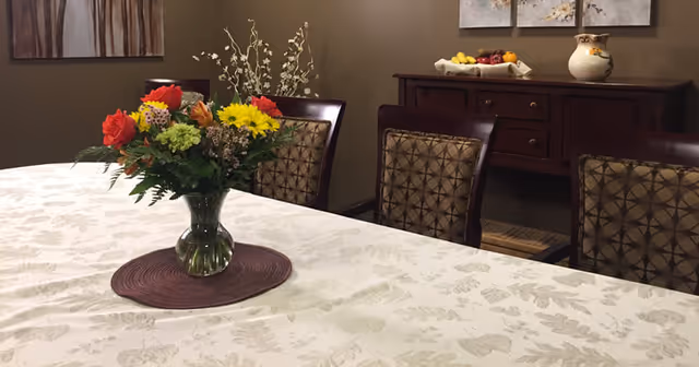 A dining room table with a white patterned tablecloth and a vase of colorful flowers in the center. Behind the table are four wooden chairs with patterned cushions. In the background, there is a wooden sideboard with a bowl of fruit and a decorative vase on top, and two pieces of wall art hanging above it.