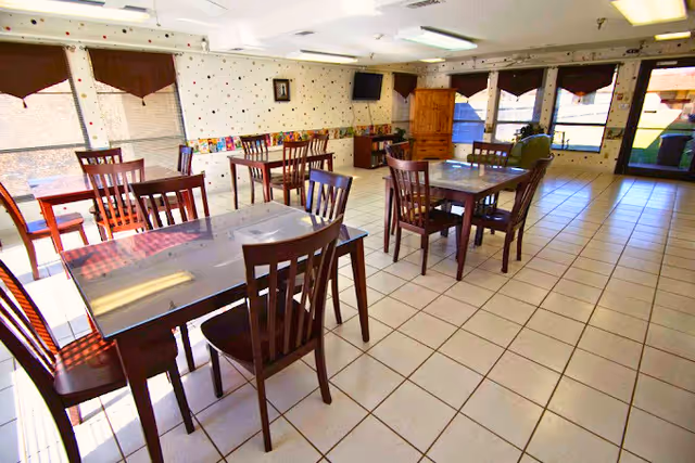 A bright dining room with several wooden tables and chairs arranged on a tiled floor. Large windows with brown valances let in natural light, and there is a wooden cabinet and a wall-mounted TV in the background. The room has a clean and simple decor with light-colored walls and a few framed pictures.