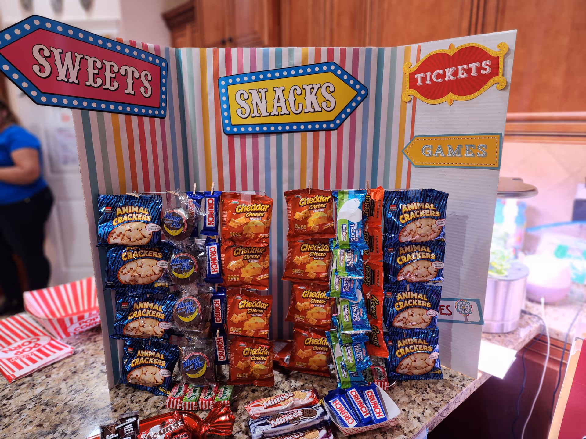 A colorful display board on a kitchen counter labeled with signs reading 'Sweets', 'Snacks', 'Tickets', and 'Games'. The board holds various packaged snacks including animal crackers, cheddar cheese crackers, Moon Pies, Crunch bars, and other candy bars. The background shows a kitchen setting with wooden cabinets and a person in a blue shirt partially visible.