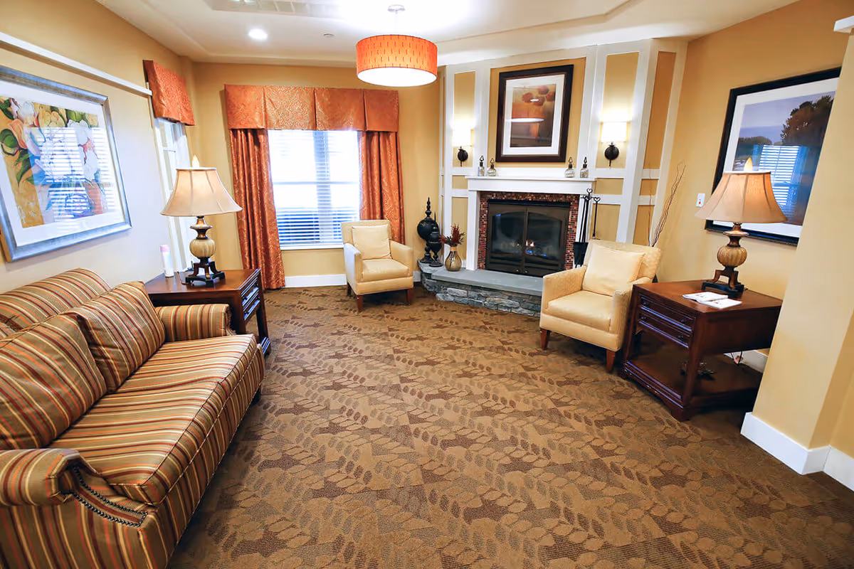A cozy living room with a striped sofa on the left, two beige armchairs facing a stone fireplace with a framed picture above it. There are two wooden side tables with lamps on either side of the room, a window with orange curtains in the background, and patterned brown carpet covering the floor.
