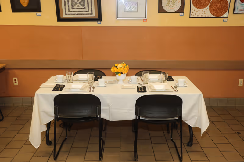 A dining table set for six people with white tablecloth, white square plates, cups, glasses, and silverware. A small vase with yellow flowers is placed in the center of the table. The table is surrounded by black chairs and is positioned against a wall decorated with framed artwork.