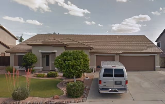Front exterior view of a single-story house with a tiled roof, a driveway with a parked silver van, and a landscaped front yard featuring green grass, bushes, and desert plants under a partly cloudy sky.