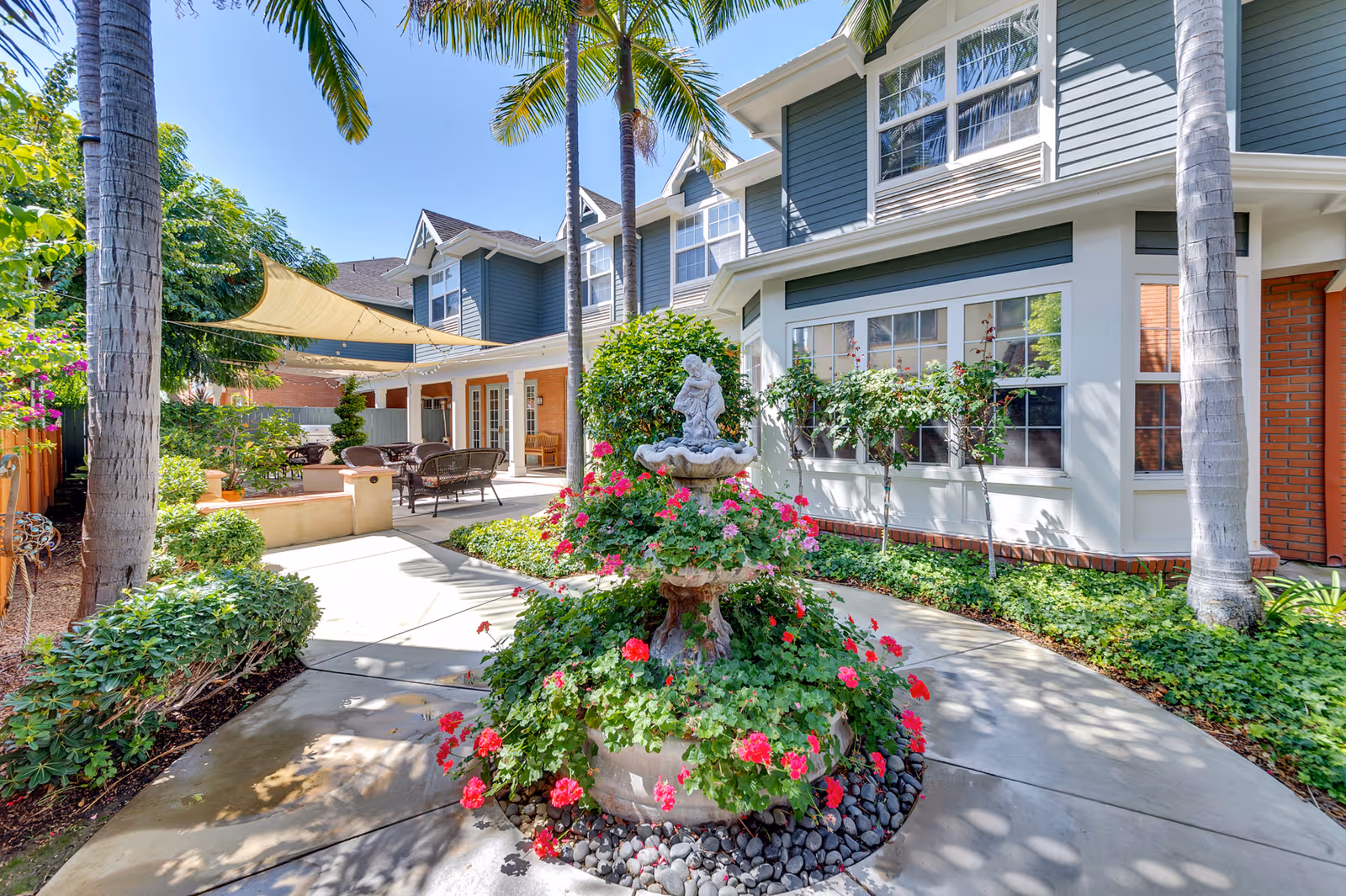 Outdoor courtyard area at Kirkwood Orange featuring a decorative tiered fountain surrounded by vibrant red and pink flowers. The courtyard is shaded by tall palm trees and beige sun sails. There are wicker chairs and tables arranged under the covered patio area attached to a blue and white building with large windows.
