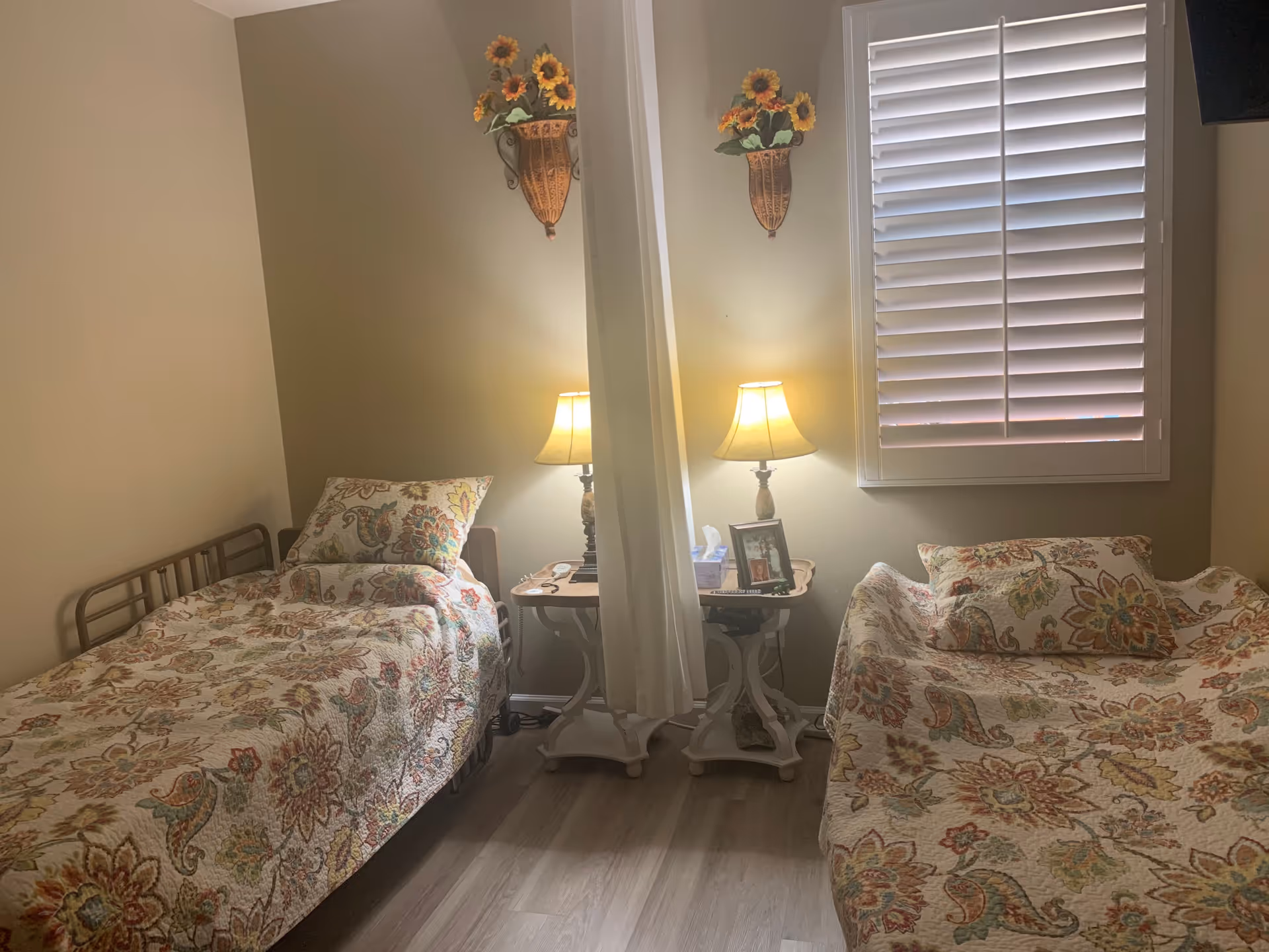 A small bedroom with two twin beds covered in floral bedspreads, a central nightstand with a lamp, and a shuttered window.