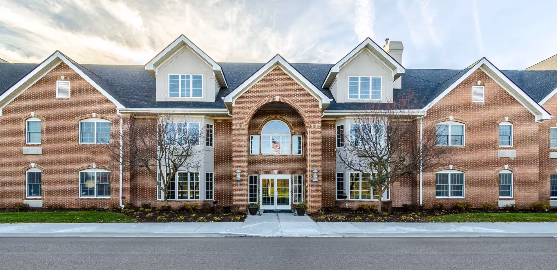 Front view of a two-story red brick assisted living building with a central arched entrance, dormer windows, and landscaped beds.