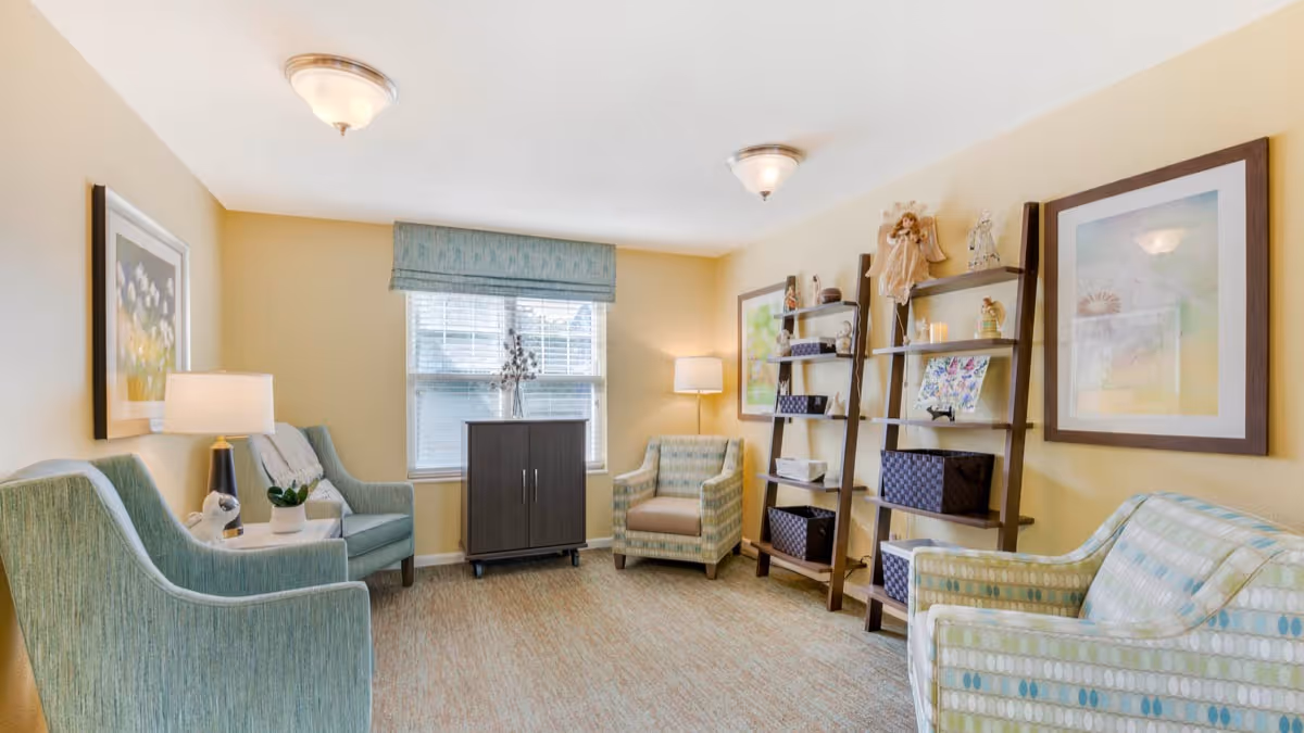 A cozy sitting room with light yellow walls, two blue-green armchairs, a patterned armchair, a small dark cabinet under a window with blinds, two wooden ladder-style shelves with decorative items, framed artwork on the walls, and two ceiling light fixtures.