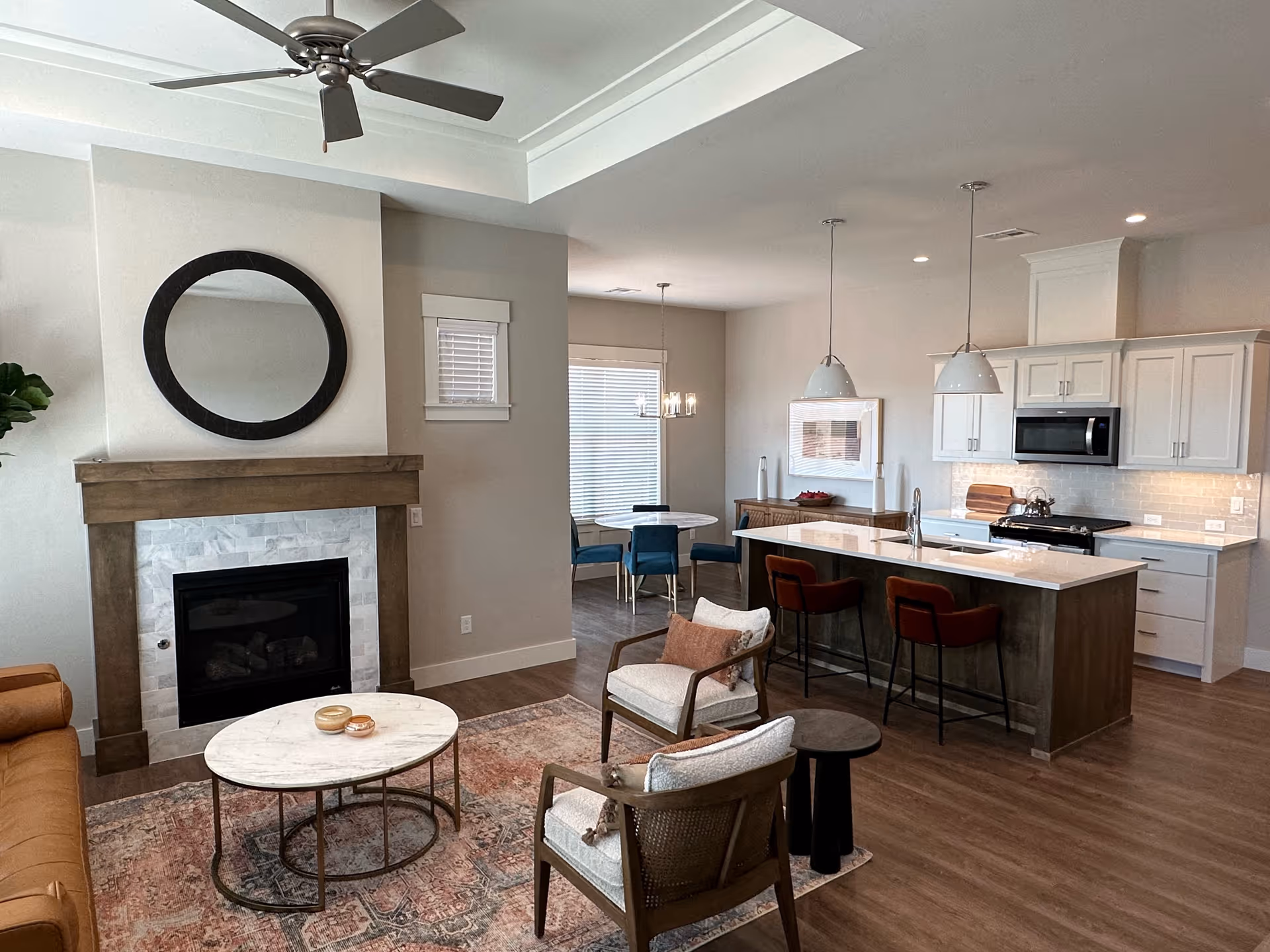 A modern open-concept living space featuring a living room with a brown leather sofa, two cushioned armchairs, a round marble coffee table, and a fireplace with a round mirror above it. Adjacent to the living room is a kitchen with a large island, two red bar stools, white cabinetry, and pendant lights. In the background, there is a dining area with a round table and blue chairs near a window with blinds.