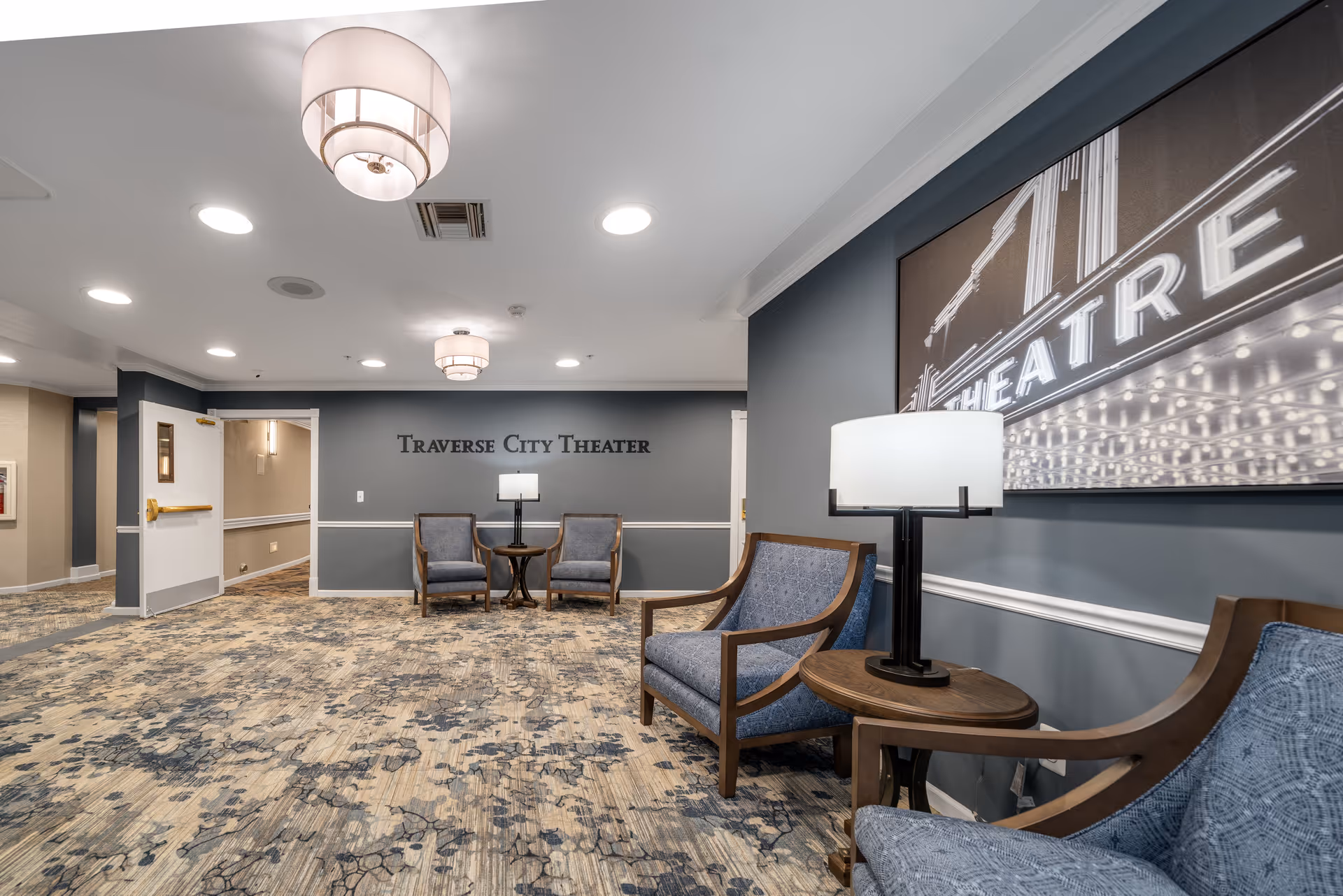 Interior view of a theater lobby area with blue-gray walls and patterned carpet. There are several blue upholstered chairs with wooden frames arranged around small wooden tables with lamps. On the wall, there is a sign that reads 'Traverse City Theater' and a large black and white photo of a theater marquee.