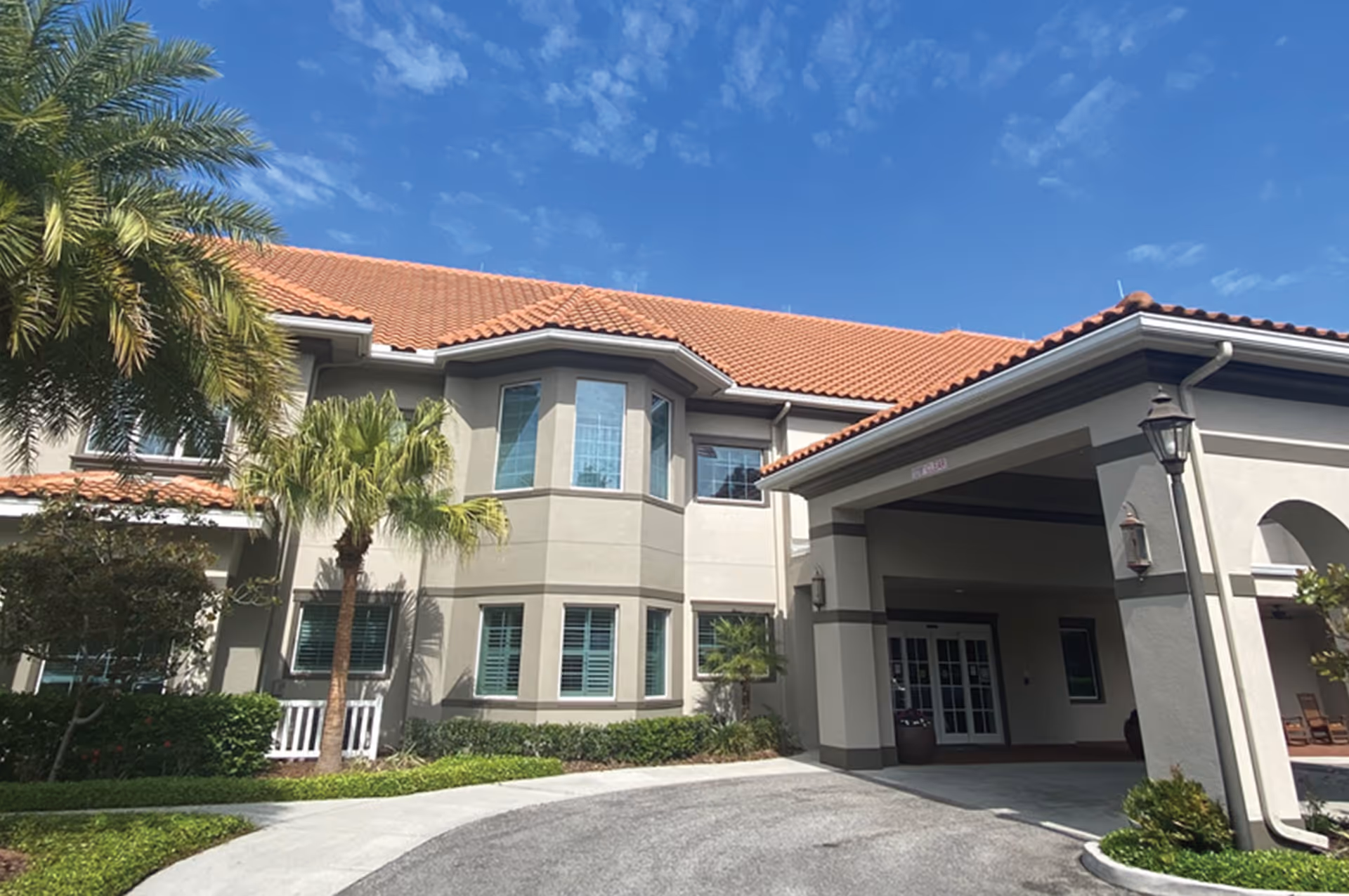 Front entrance of a two-story Mediterranean-style building with a red tile roof, palm trees, and a covered porte-cochère.