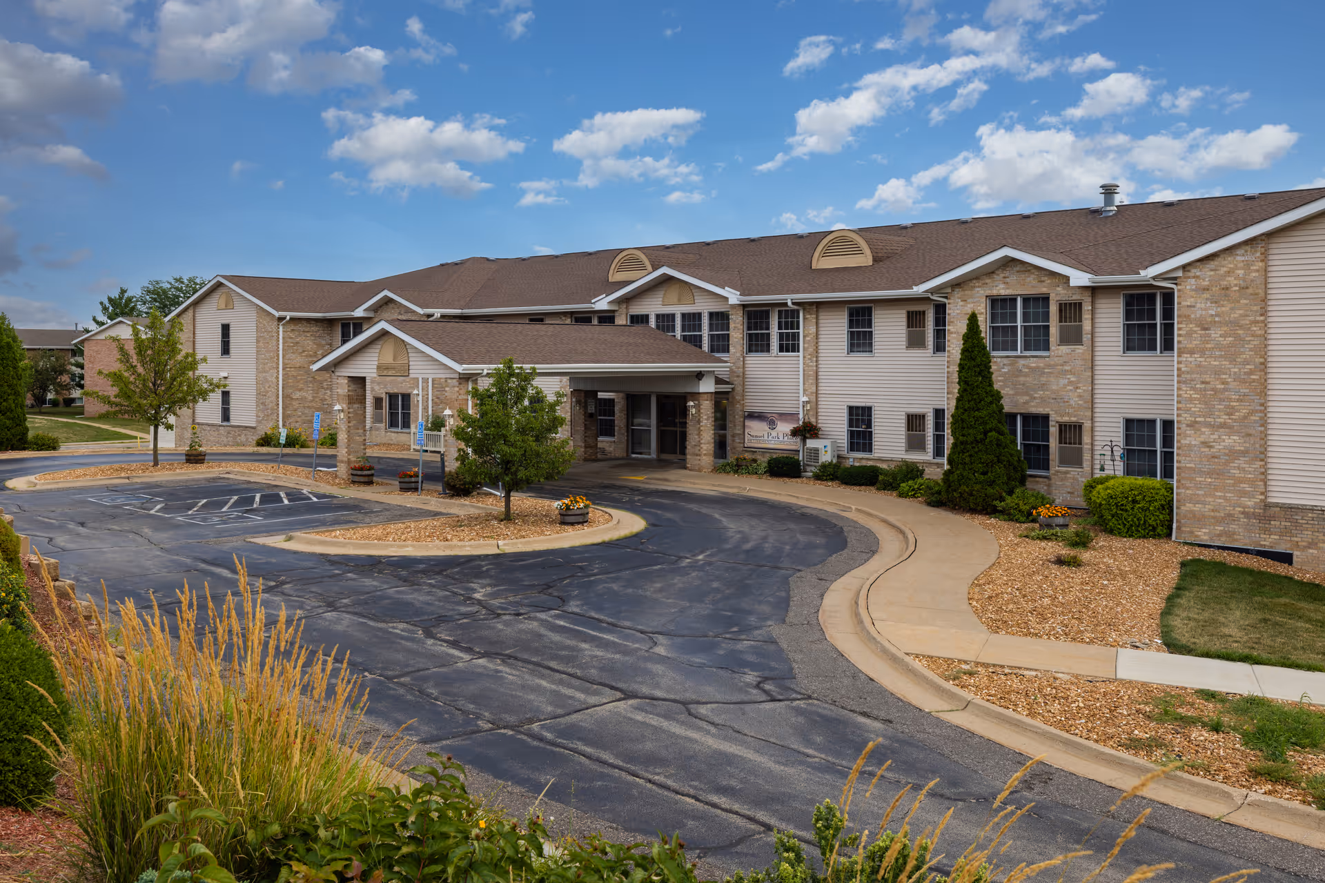 Exterior view of Sunset Park Place, a two-story senior living facility with a covered entrance, surrounded by landscaped greenery and a paved driveway under a partly cloudy sky.