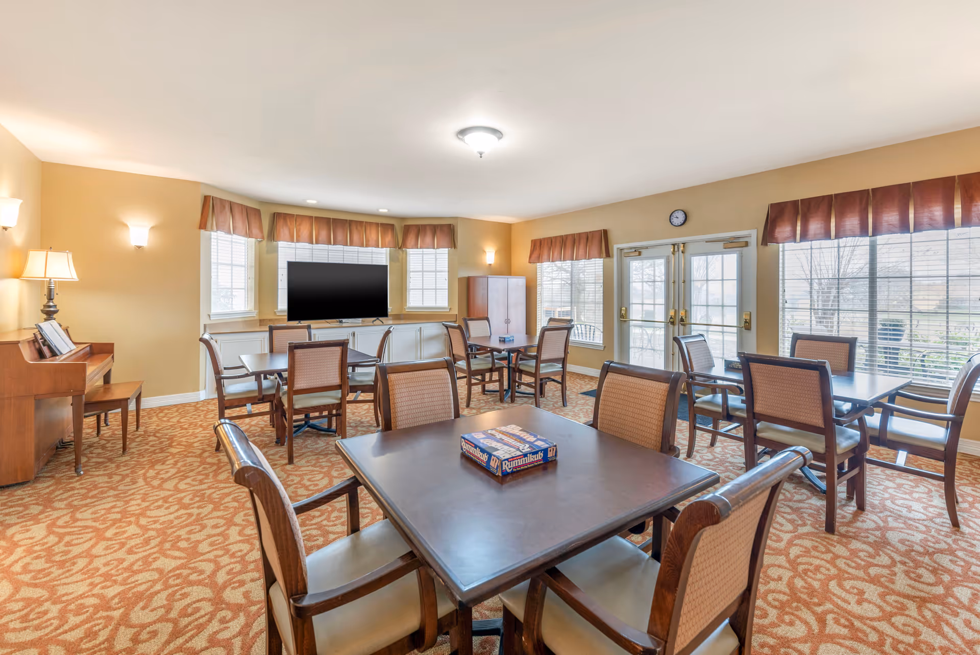 A well-lit common room with multiple square tables and chairs arranged for group activities or dining. A box of the game Rummikub is placed on the nearest table. The room has large windows with brown valances, a piano with a lamp on the left side, a flat-screen TV mounted on a cabinet, and double glass doors leading outside.