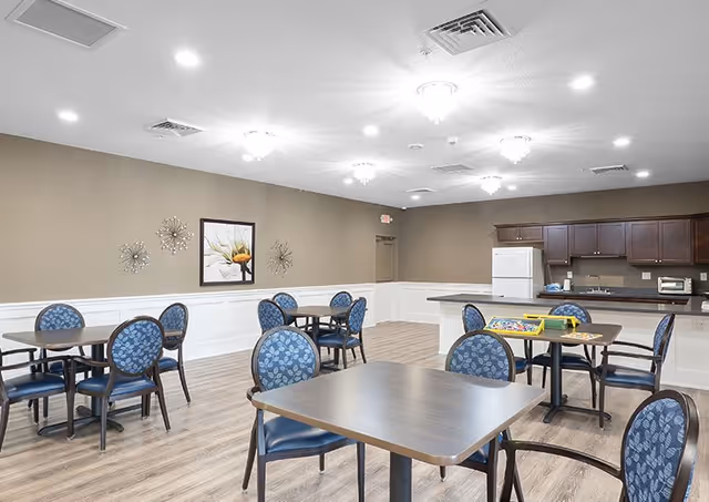 Bright communal dining/activity room with several tables and blue patterned chairs and a kitchenette along the back wall.