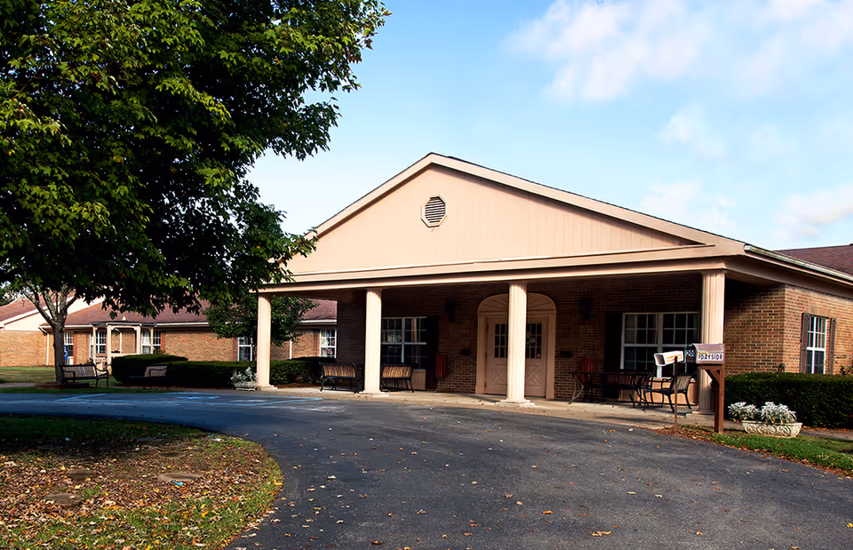 Exterior view of Braxton Healthcare Center showing a single-story brick building with a covered entrance supported by columns. There are benches on the porch and a tree with green leaves on the left side. The driveway curves in front of the entrance under a partly cloudy sky.