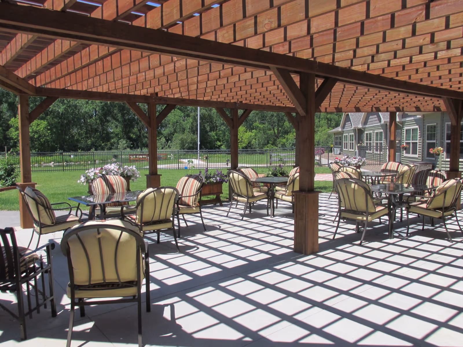Outdoor seating area with cushioned chairs and glass-top tables under a wooden pergola casting patterned shadows on the ground. In the background, there is a green lawn, trees, and a building with windows and flower pots.