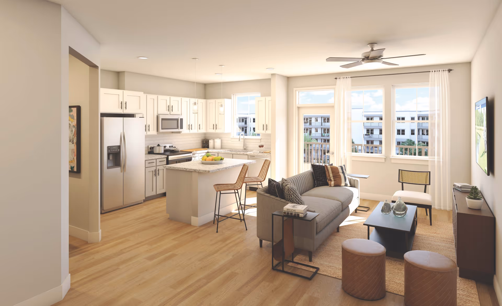 A bright and modern open-concept living room and kitchen area with light wood flooring. The kitchen features white cabinets, stainless steel appliances including a refrigerator and microwave, and a central island with two wicker bar stools. The living room has a gray sofa with multiple pillows, a coffee table with decorative glass bottles, two round ottomans, a chair, and a wall-mounted TV. Large windows and a glass door let in natural light and offer a view of neighboring buildings.