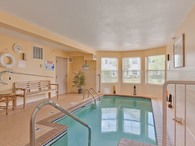 Indoor community swimming pool in a recreation room with tiled deck, handrails, bench seating, and windows.