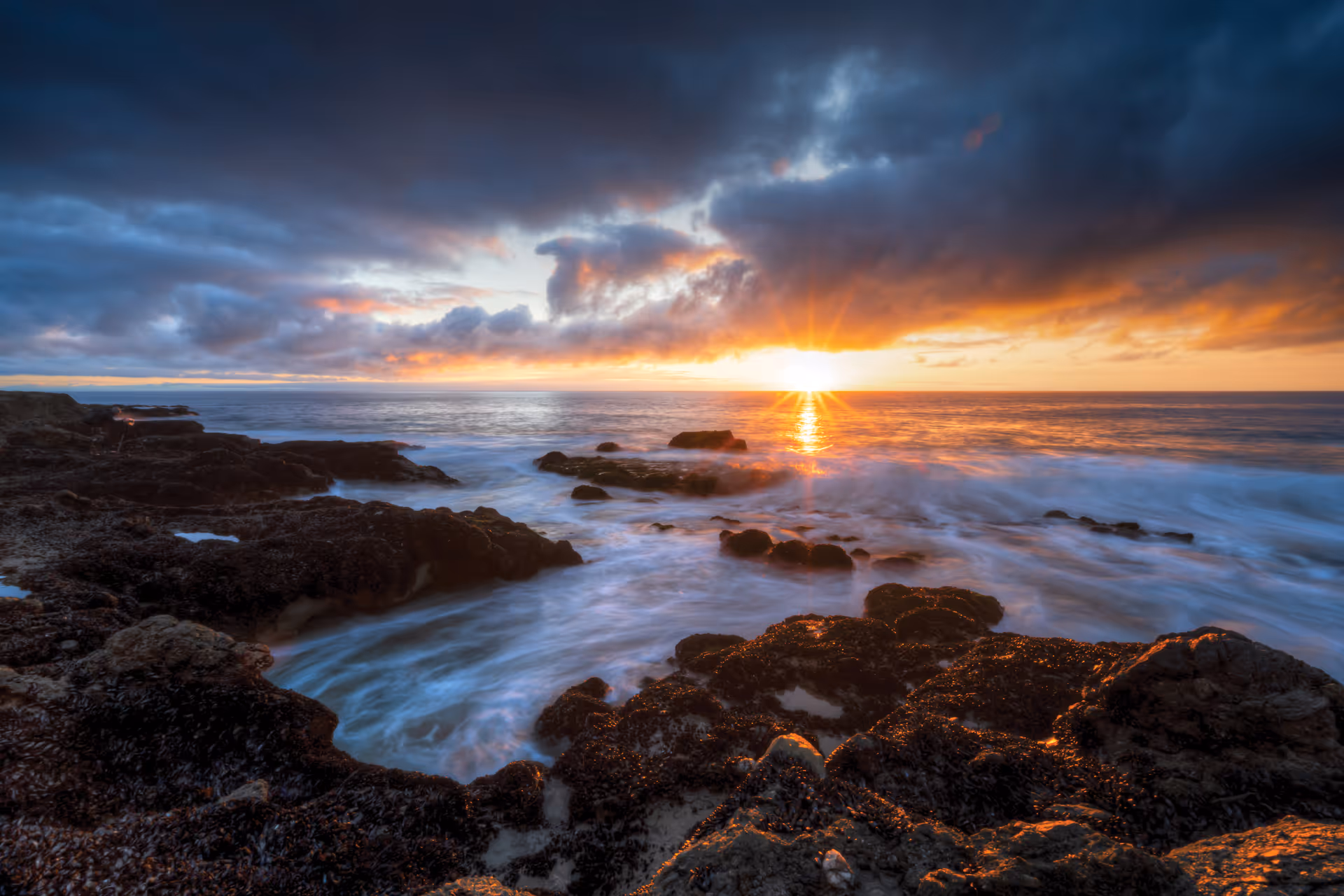 Sunset over a rocky shoreline with waves and dramatic clouds.