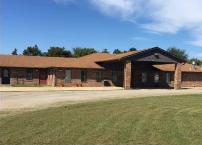 Single-story brick building with a brown shingled roof under a blue sky, featuring a covered entrance and several windows, surrounded by a grassy area and a dirt driveway.