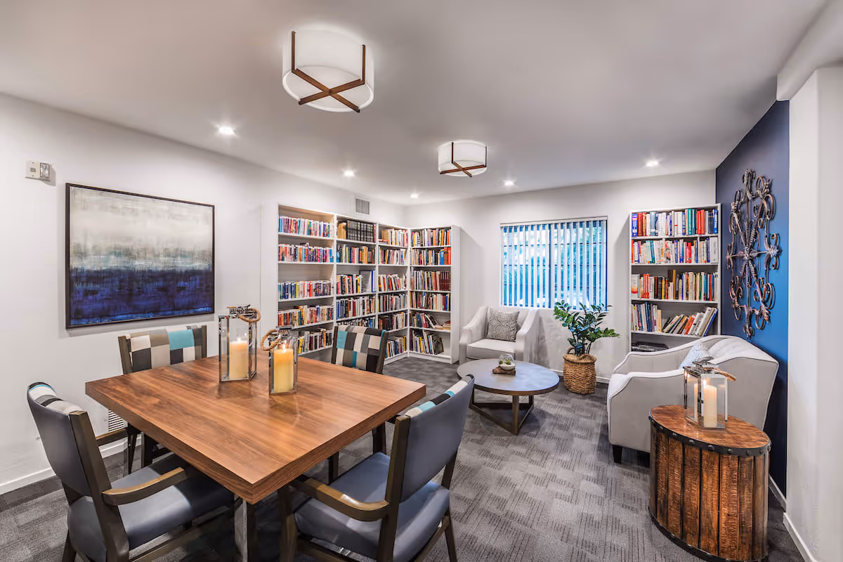 A cozy library and sitting area with white walls and carpeted floor. The room features a wooden table with four chairs, two large candles in glass holders on the table, multiple bookshelves filled with books, a white armchair, a white sofa, a round wooden coffee table, a potted plant, and decorative wall art including a large abstract painting and a metal wall sculpture. Vertical blinds cover the window, allowing natural light to enter.