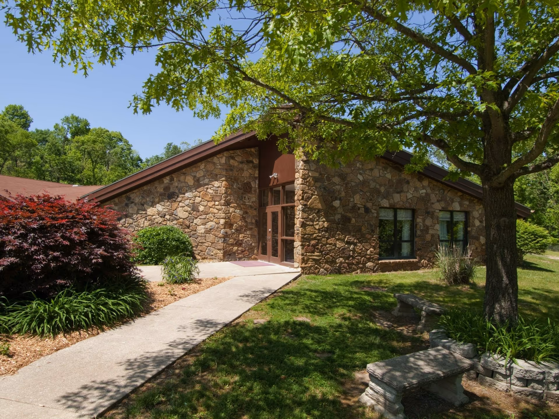 Stone-faced single-story building entrance with a concrete walkway, landscaped shrubs, benches, and trees in front.