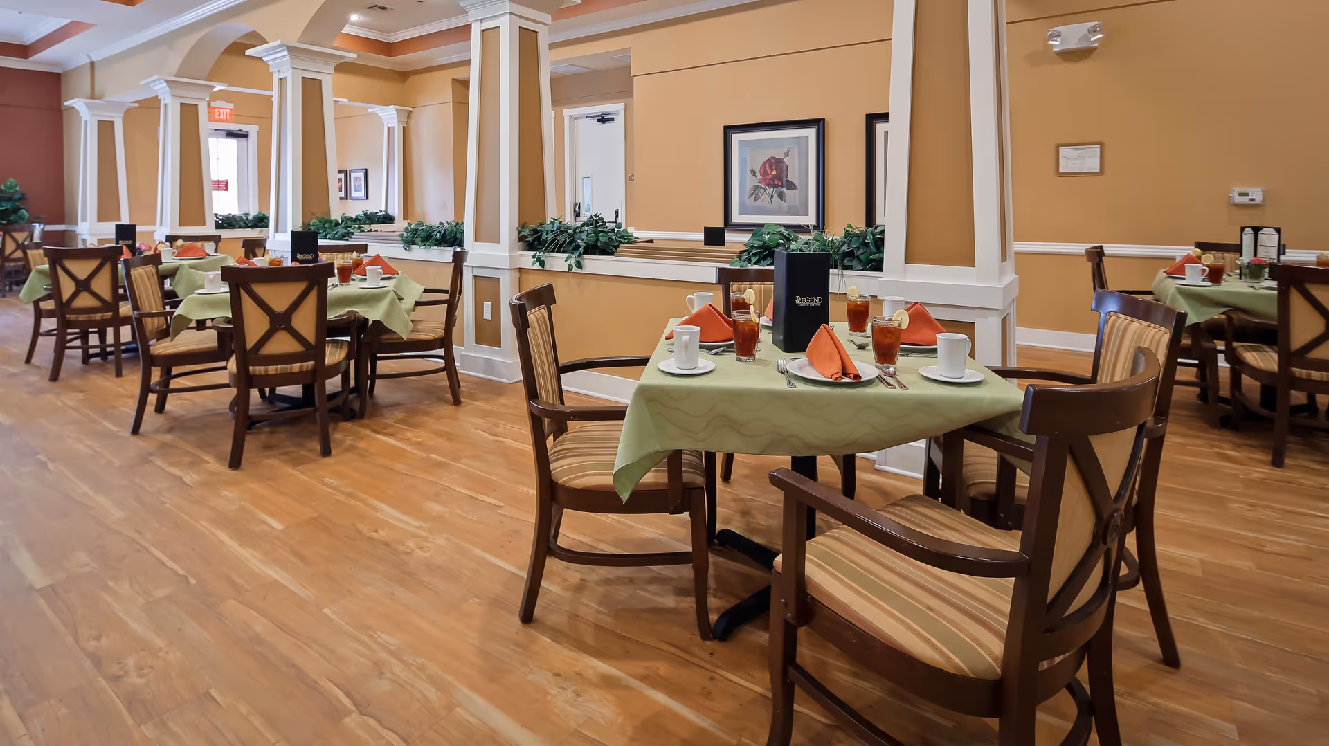A dining room in an assisted living facility with several tables covered with light green tablecloths, each set with orange napkins, cups, glasses of iced tea, and menus. The room has wooden floors, beige walls, white columns, and decorative plants on ledges.