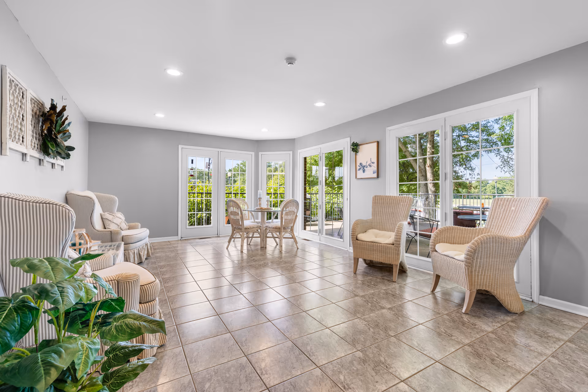 Sunlit common room with wicker armchairs, a small dining table, tiled floor, and large glass doors/windows opening to outdoor greenery.