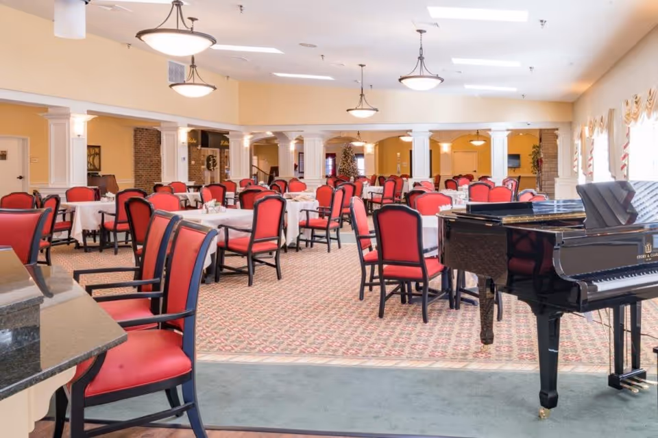 A spacious dining room with multiple tables covered in white tablecloths and surrounded by red cushioned chairs. The room features a patterned carpet, cream-colored walls, large windows with curtains, and several ceiling lights. A black grand piano is positioned on the right side of the image.