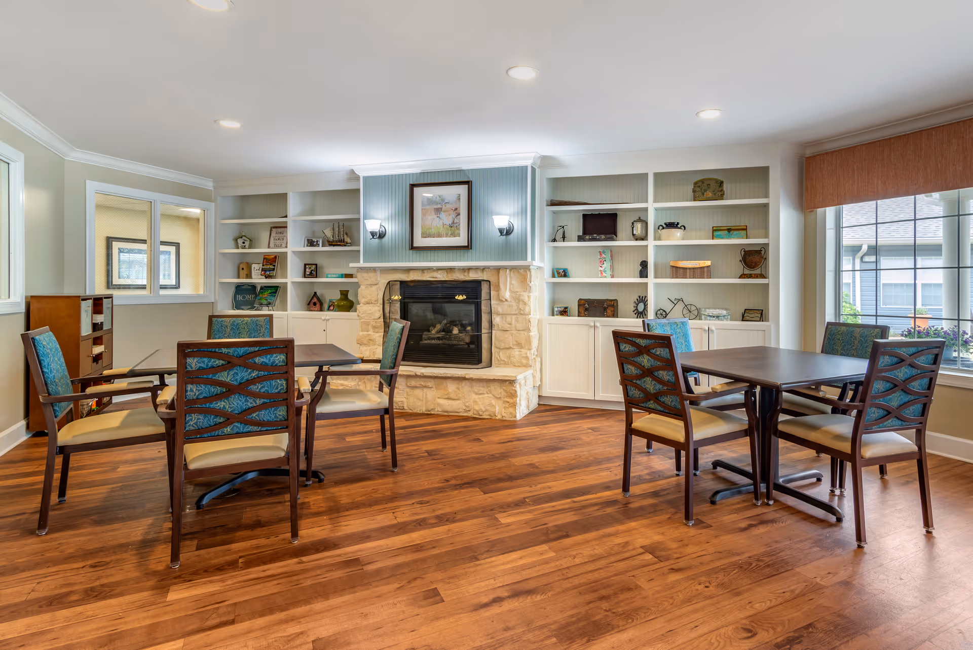 A cozy common area with wooden flooring, two tables each surrounded by four chairs with blue patterned backs and beige seats, a stone fireplace in the center with a framed picture above it, and white built-in shelves on either side displaying decorative items. Large windows on the right side let in natural light.